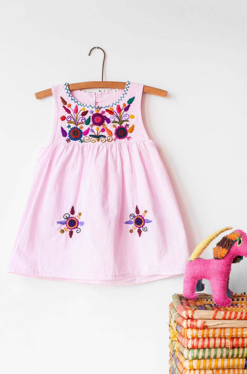 Kids sleeveless light pinki sun dress hanging with multicolor hand-embroidered floral pattern on chest and two hand-embroidered flowers near the bottom of the skirt on the front left and right.