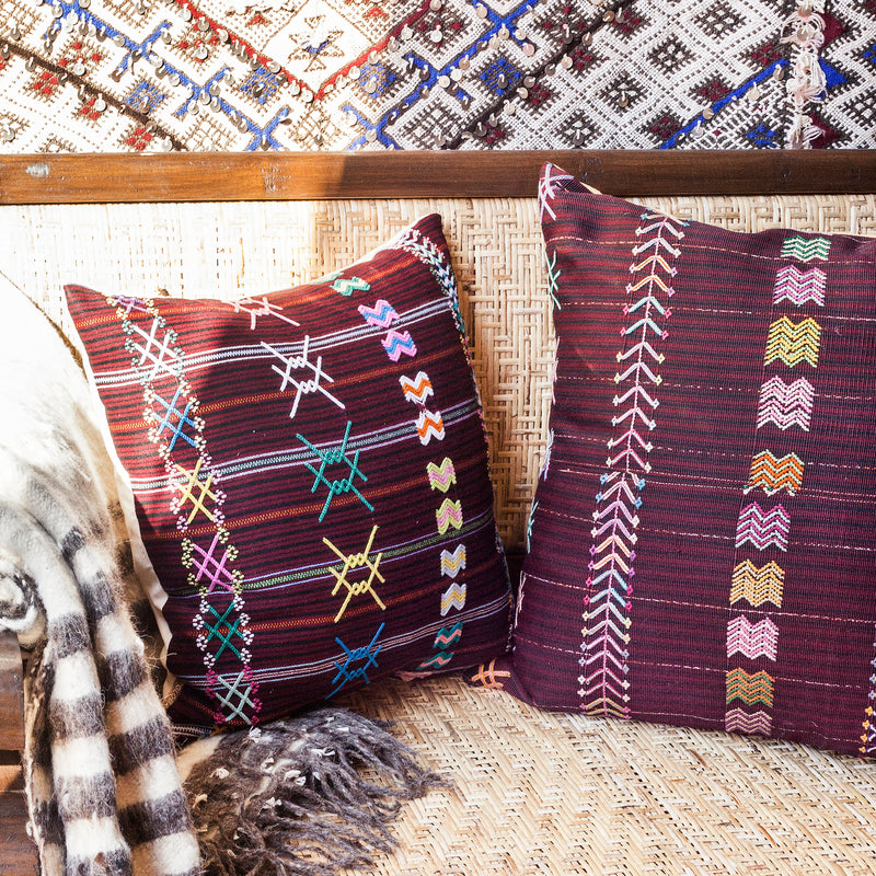 A pair of square throw pillows with a striped brown burgundy textile and colorful hand embroidered brocade on a couch