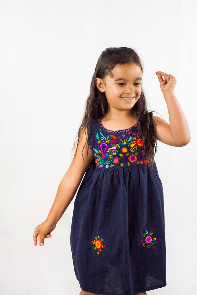Child wearing a sleeveless navy knee-length sun dress. There are multicolor hand embroidered floral elements on the chest and skirt of the dress and cross stitching at the neckline.