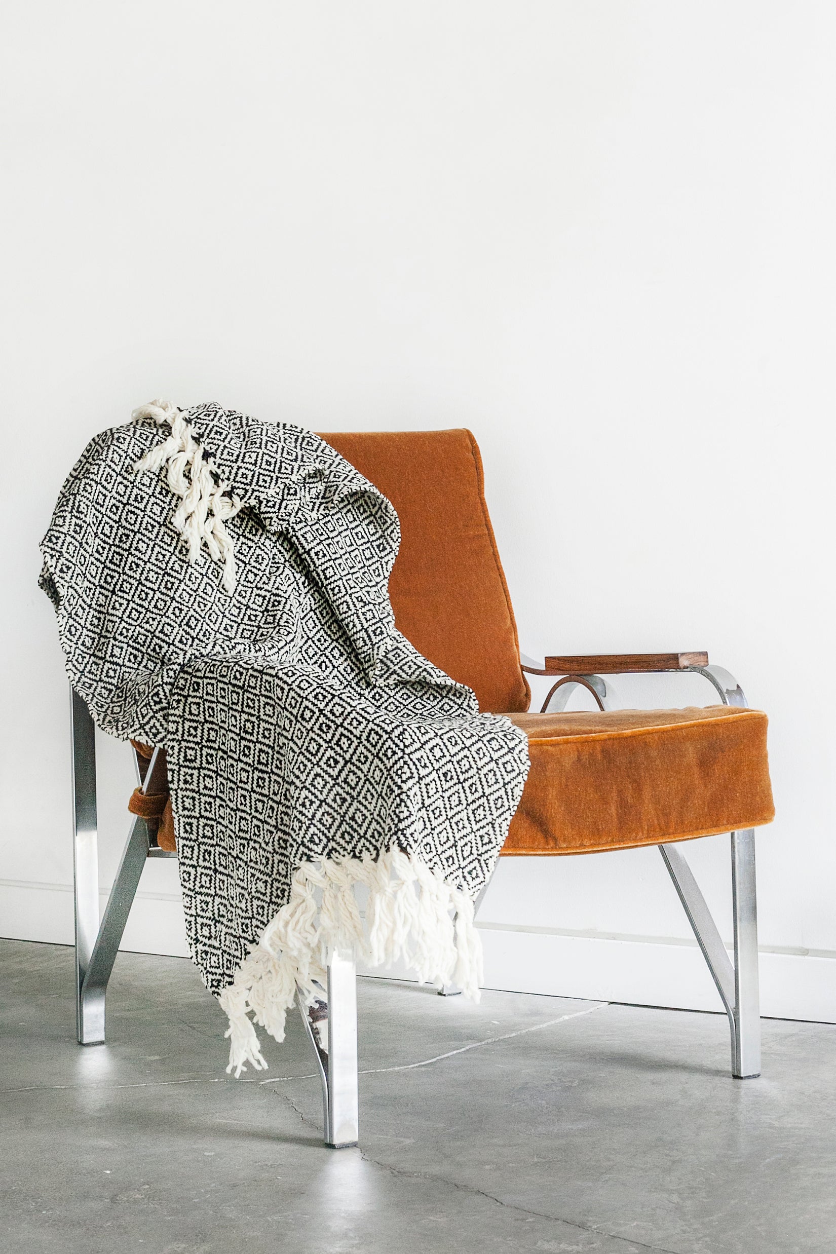 Black and white wool throw blanket woven in a diamond pattern with hand tied tassels at each end draped over a mid century modern chair