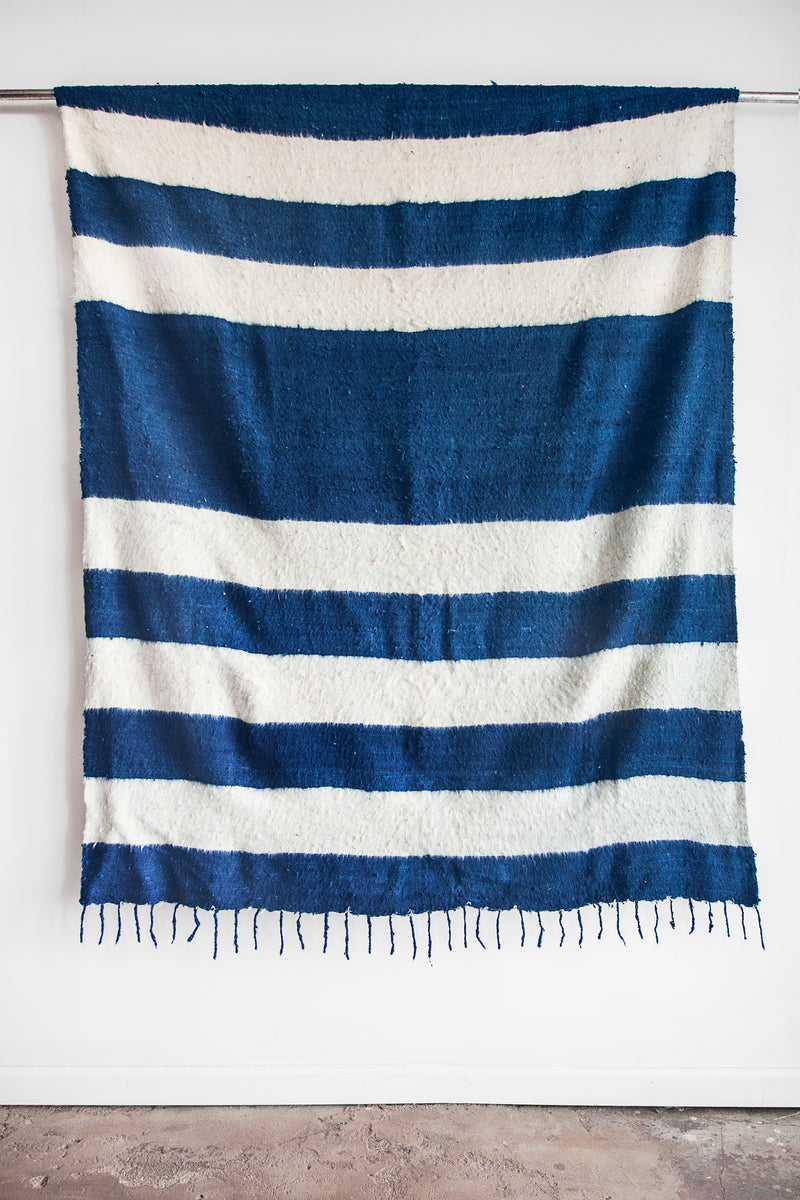 Queen sized ecru wool blanket with rows of thick horizontal blue indigo stripes and a row of tied tassels at each end
