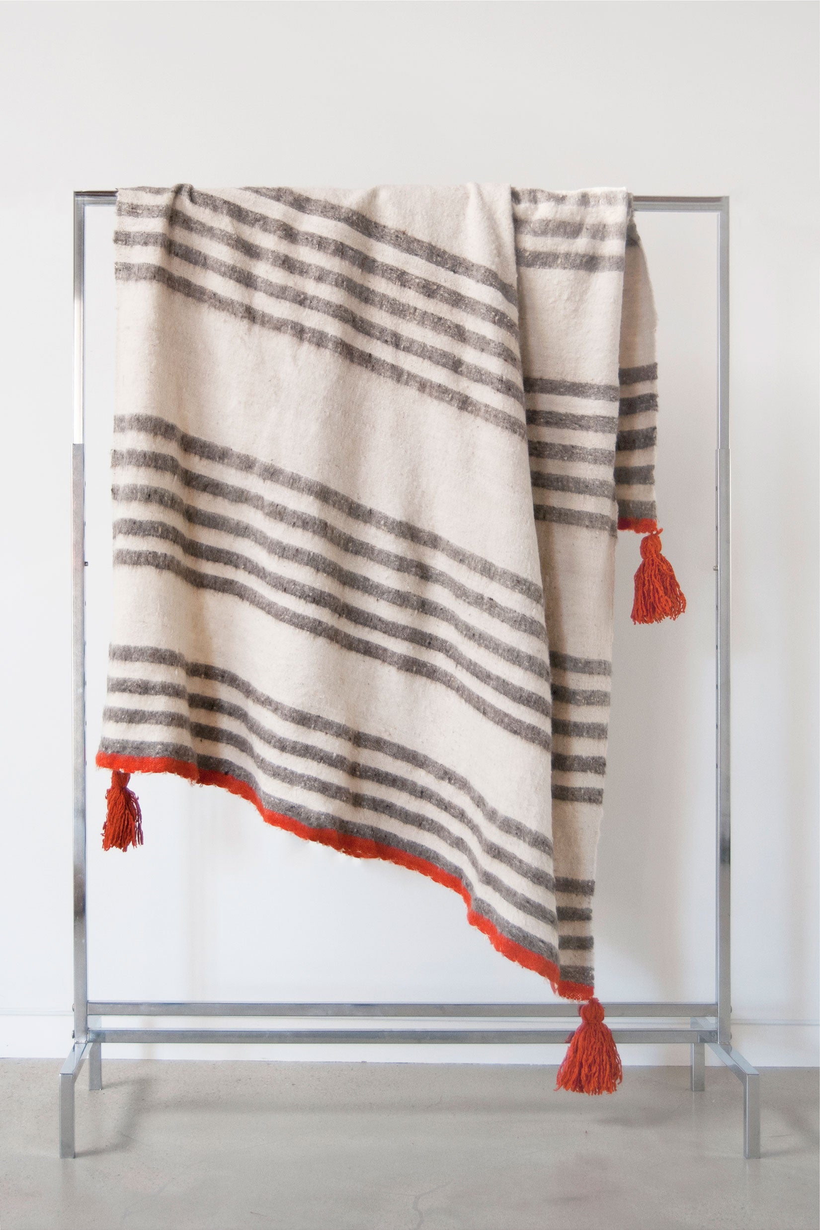 Queen size ecru wool blanket with rows of thin grey stripes throughout and pumpkin accent stripes at both ends and pumpkin tassels at each corner
