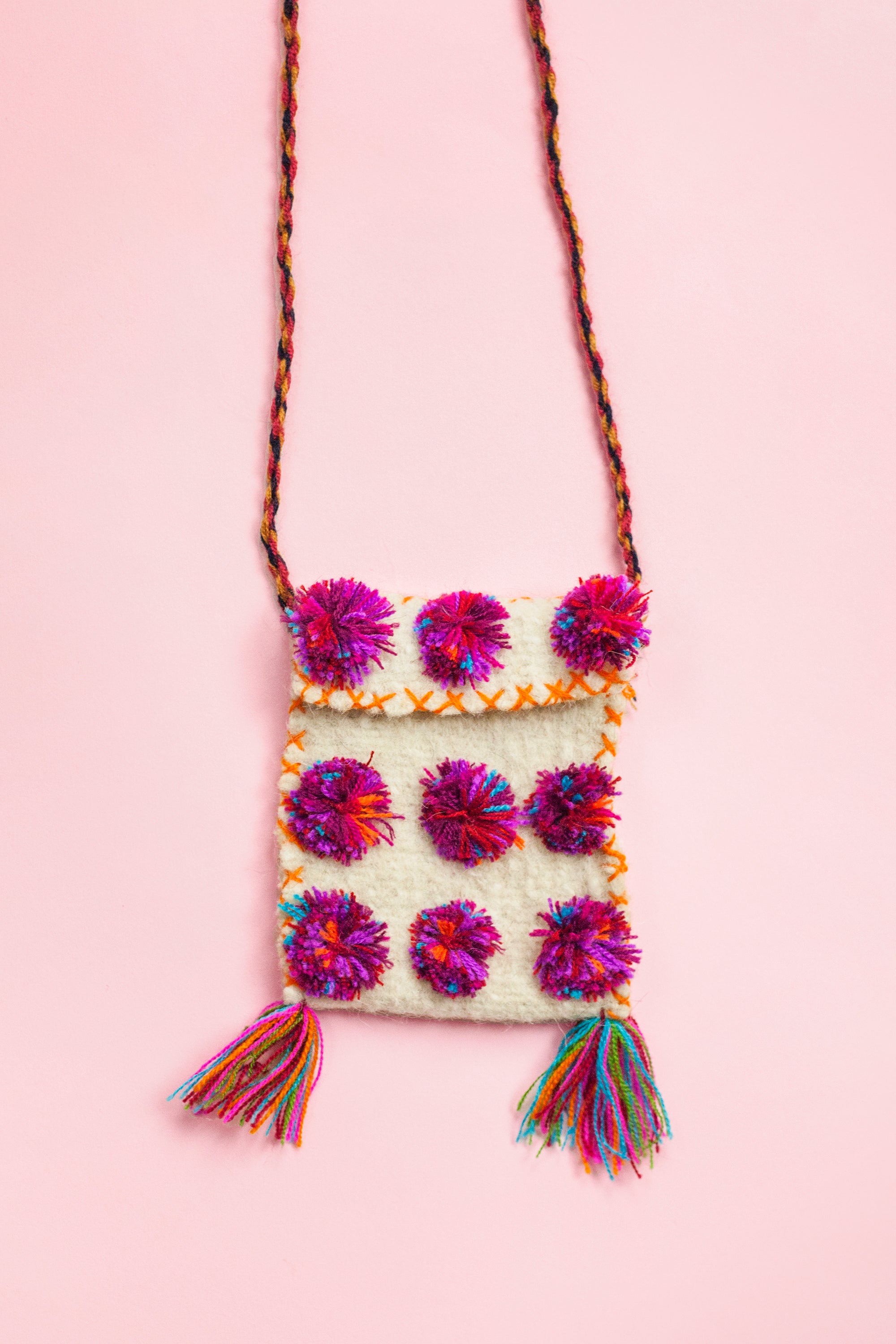White wool satchel covered in three rows of multicolor pom poms. There is colorful cross stitching around the bag body, two small multicolor fringe tassels at the bottom corners, and a multicolor braided strap holding it up.