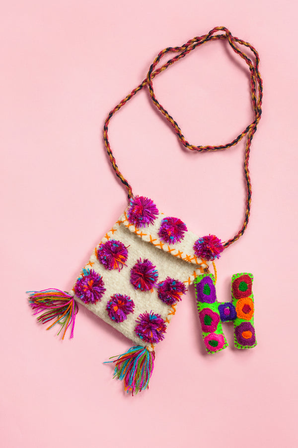 White wool satchel covered in three rows of multicolor pom poms. Along the bag there is colorful cross stitching, two small multicolor fringe tassels at the bottom corners, and a multicolor braided strap holding it. A small plush felt letter "H" embroidered with multicolor flowers is sitting next to it.