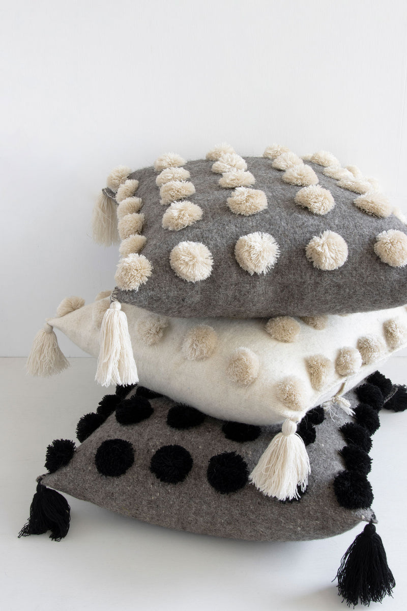 Three square woven wool throw pillows in different neutral colors covered in pom poms with tassels in each corner stacked on top of each other