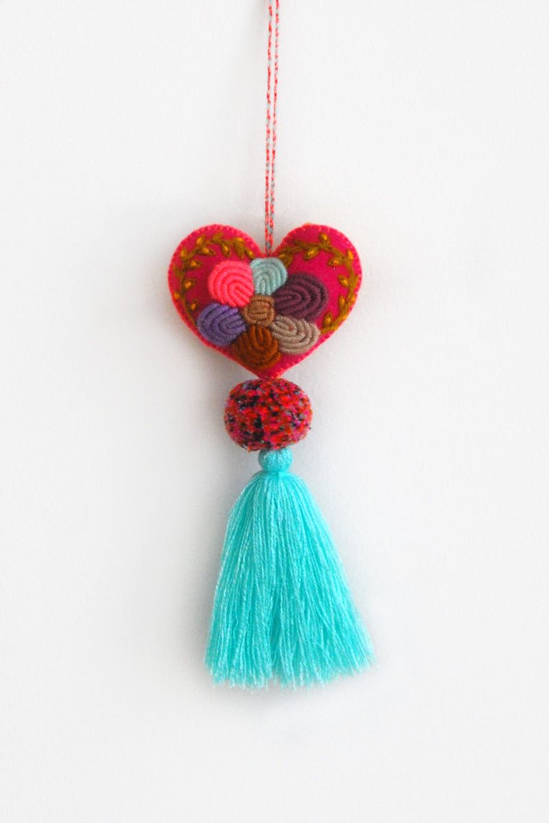 A colorful plush felt heart adorned with flower embroidery attached by multicolor speckled pom poms to a tassel in a bright blue color. The heart is hanging from a string attached to the top of it.