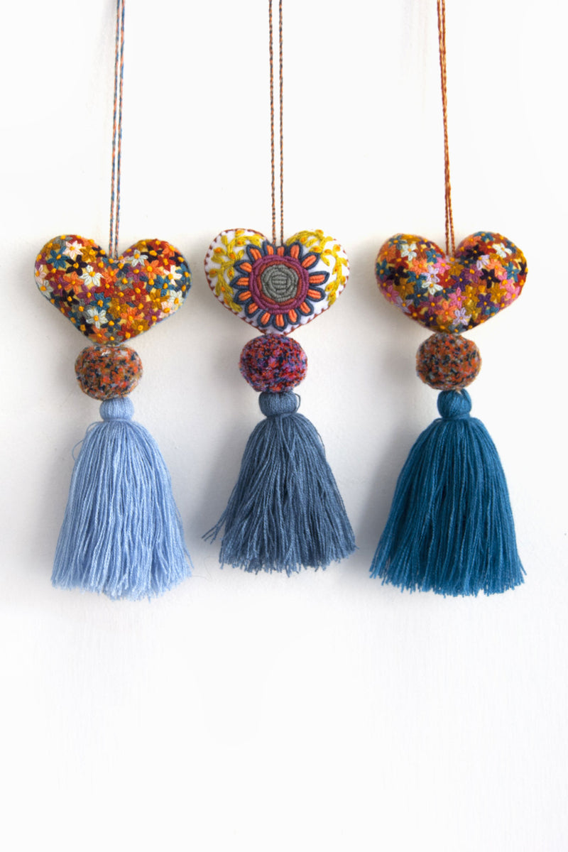 Three colorful plush felt hearts adorned with flower embroidery attached by multicolor speckled pom poms to tassels in varying shades of blue. Each heart is hanging from a string attached to the top of it.