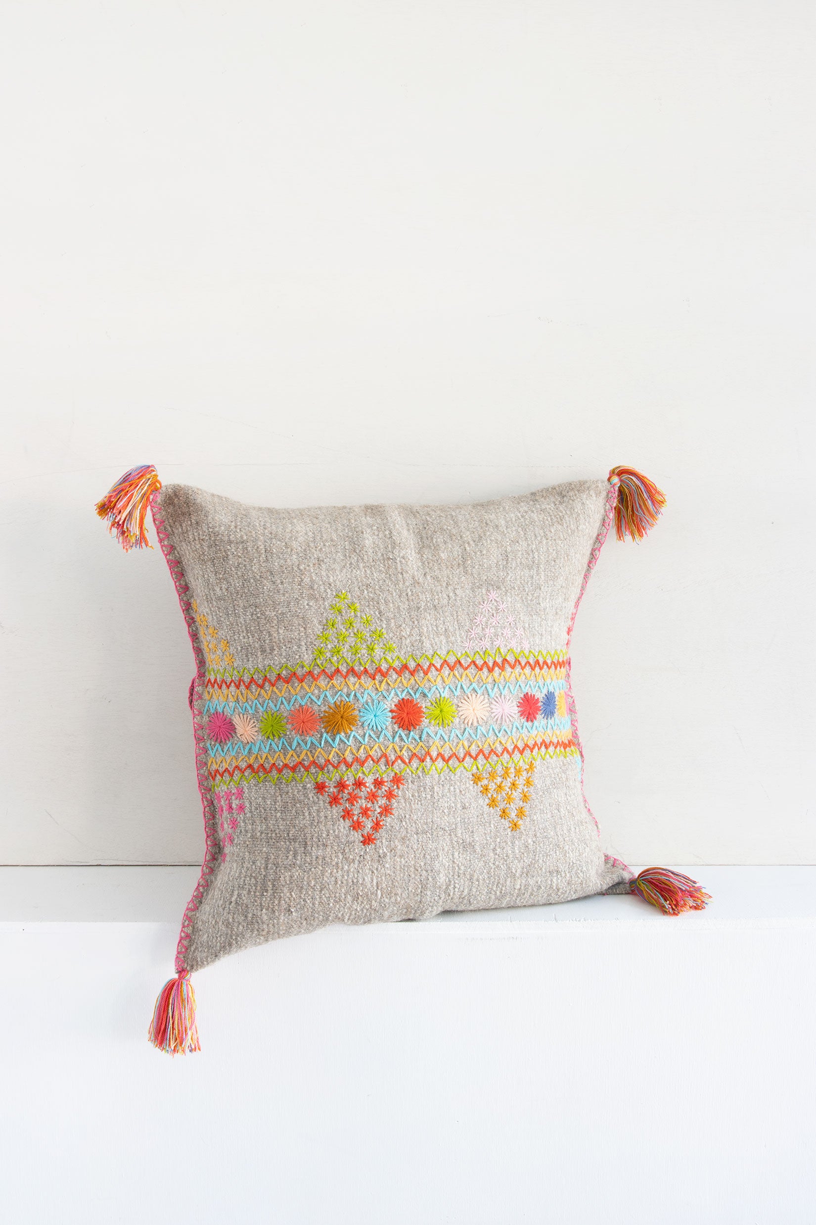 Square woven light grey wool throw pillow with multicolor star and zig-zag embroidery and tassels at each corner