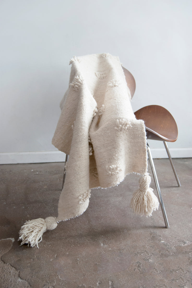 Cream wool runner with staggered rows of fringe accents throughout and cream tassels at each corner draped over a chair