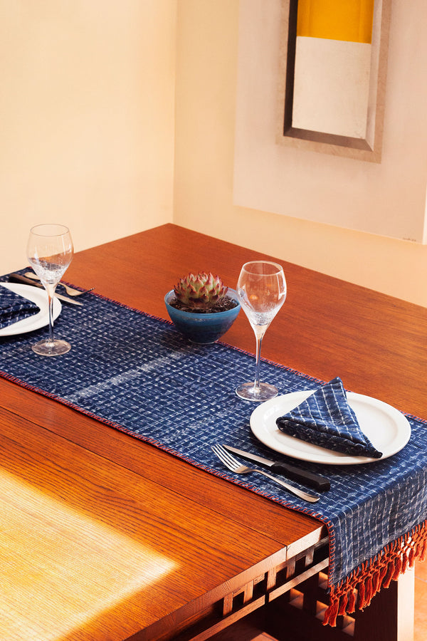 Wood table set with a blue and white patterned runner, a potted succulent, two wine glasses, silverware, plates, and blue and white patterned napkins folded on the plates.