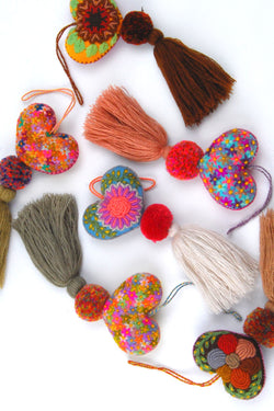 A scatter of colorful plush felt hearts adorned with flower embroidery attached by multicolor speckled pom poms to tassels in neutral shades. Each heart has a hanging string attached to the top of it.