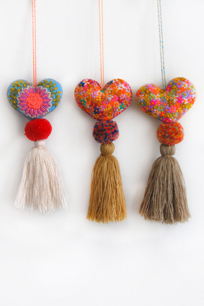 Colorful plush felt hearts adorned with flower embroidery attached by multicolor speckled pom poms to tassels in neutral shades. Each heart has a hanging string attached to the top of it.