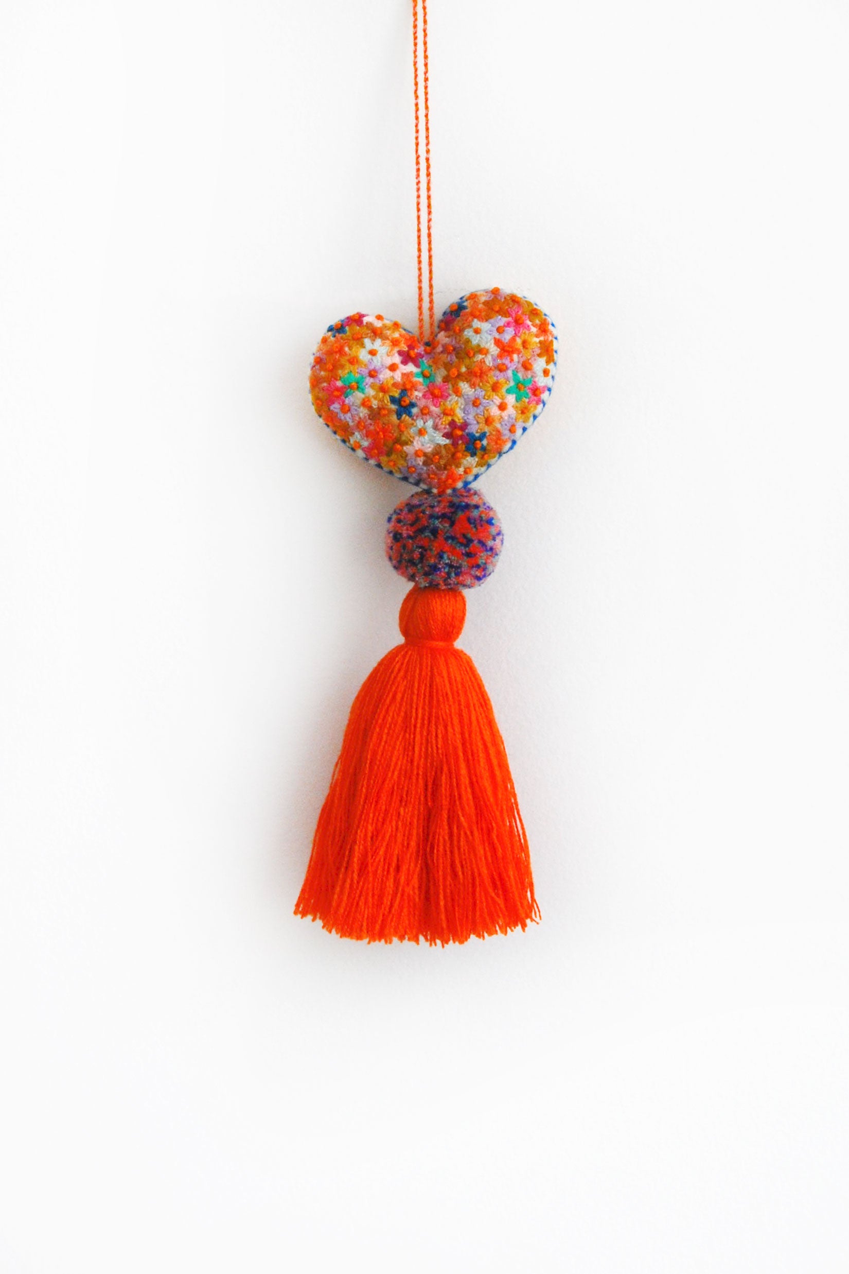 A colorful plush felt heart adorned with flower embroidery attached by multicolor speckled pom poms to a tassel in a bright orange color. The heart is hanging from a string attached to the top of it.
