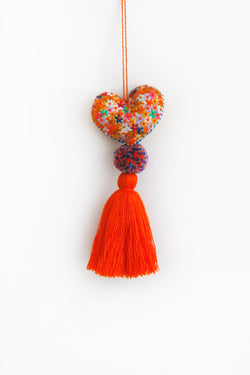 A colorful plush felt heart adorned with flower embroidery attached by multicolor speckled pom poms to a tassel in a bright orange color. The heart is hanging from a string attached to the top of it.