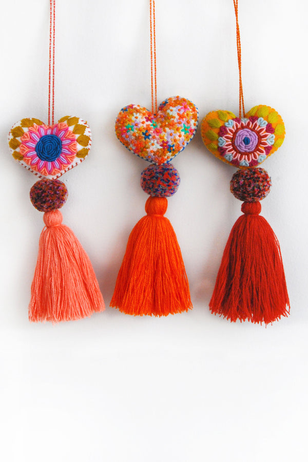 Three colorful plush felt hearts adorned with flower embroidery attached by multicolor speckled pom poms to tassels in varying shades of orange. Each heart is hanging from a string attached to the top of it.