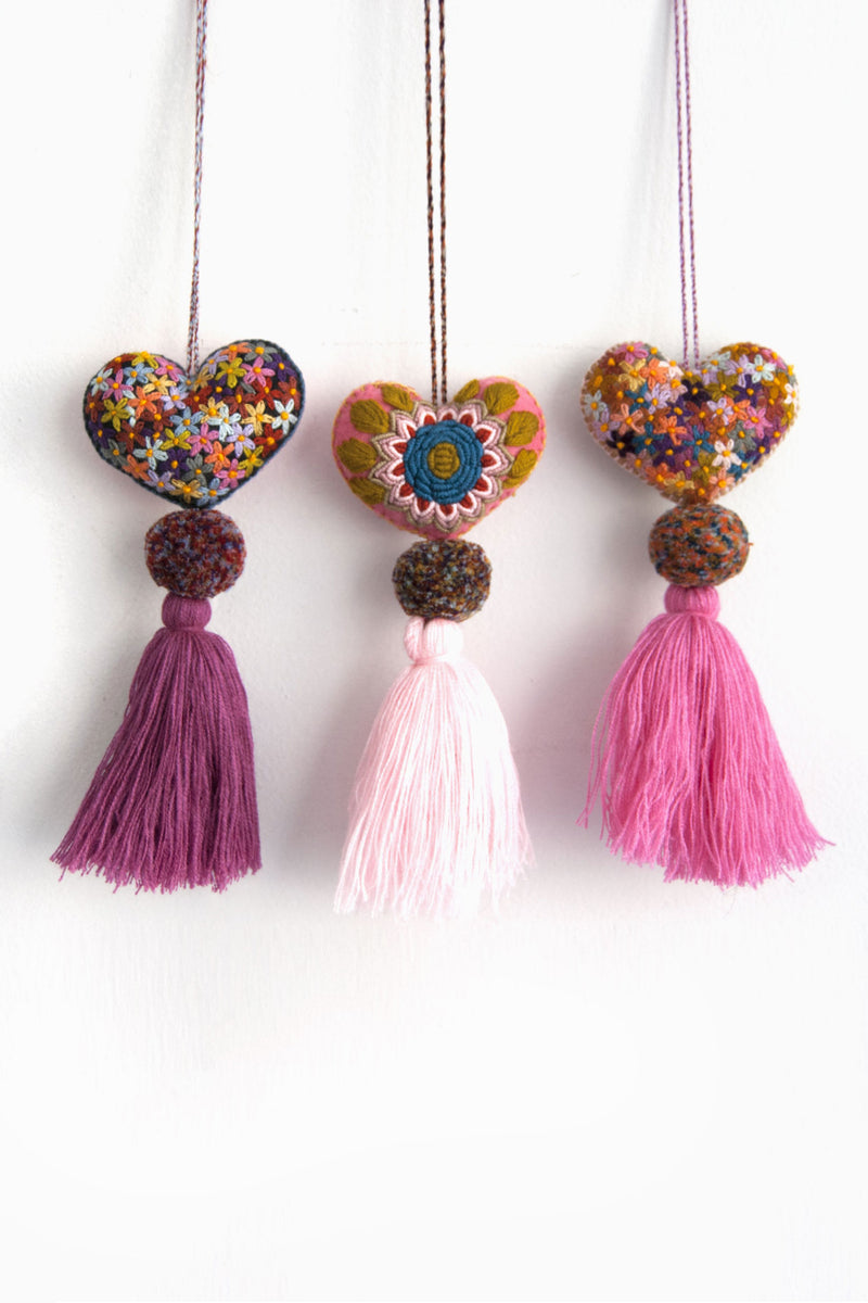 Three colorful plush felt hearts adorned with flower embroidery attached by multicolor speckled pom poms to tassels in varying shades of pink. Each heart is hanging from a string attached to the top of it.
