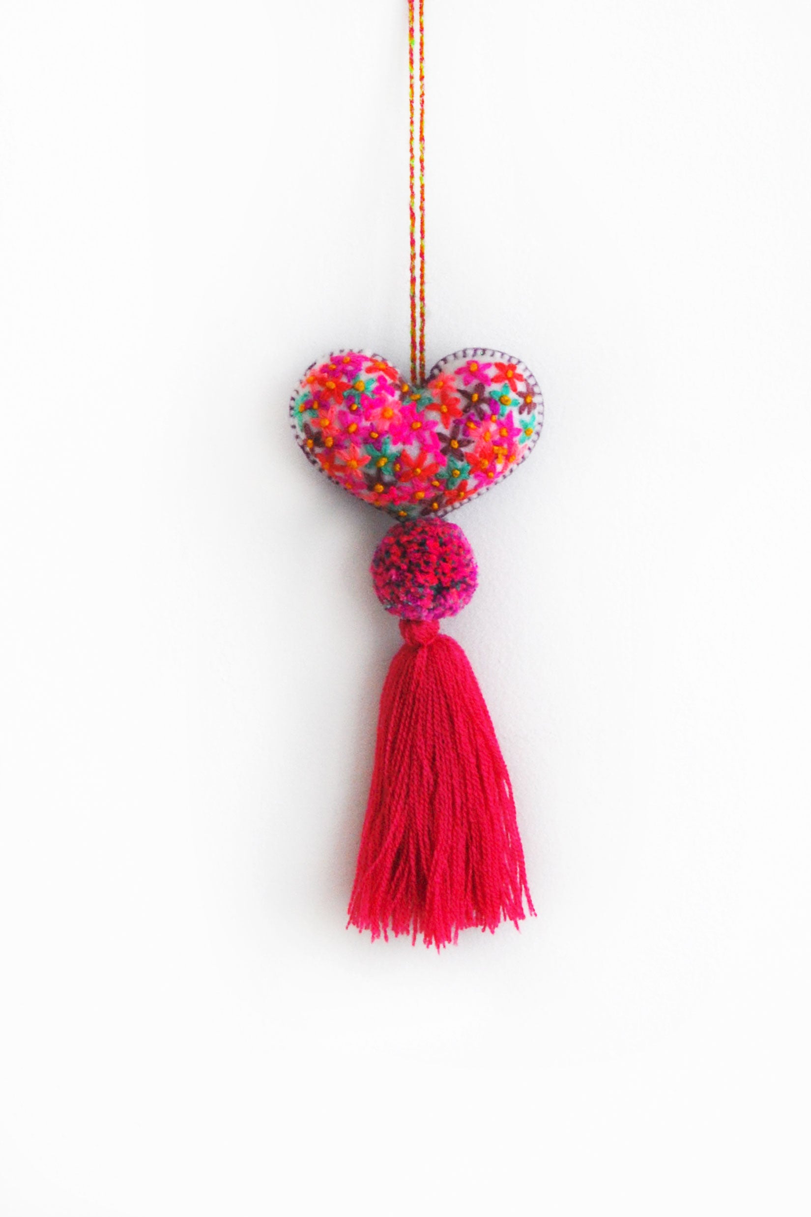 A colorful plush felt heart adorned with flower embroidery attached by multicolor speckled pom poms to a tassel in a bright pink color. The heart is hanging from a string attached to the top of it.