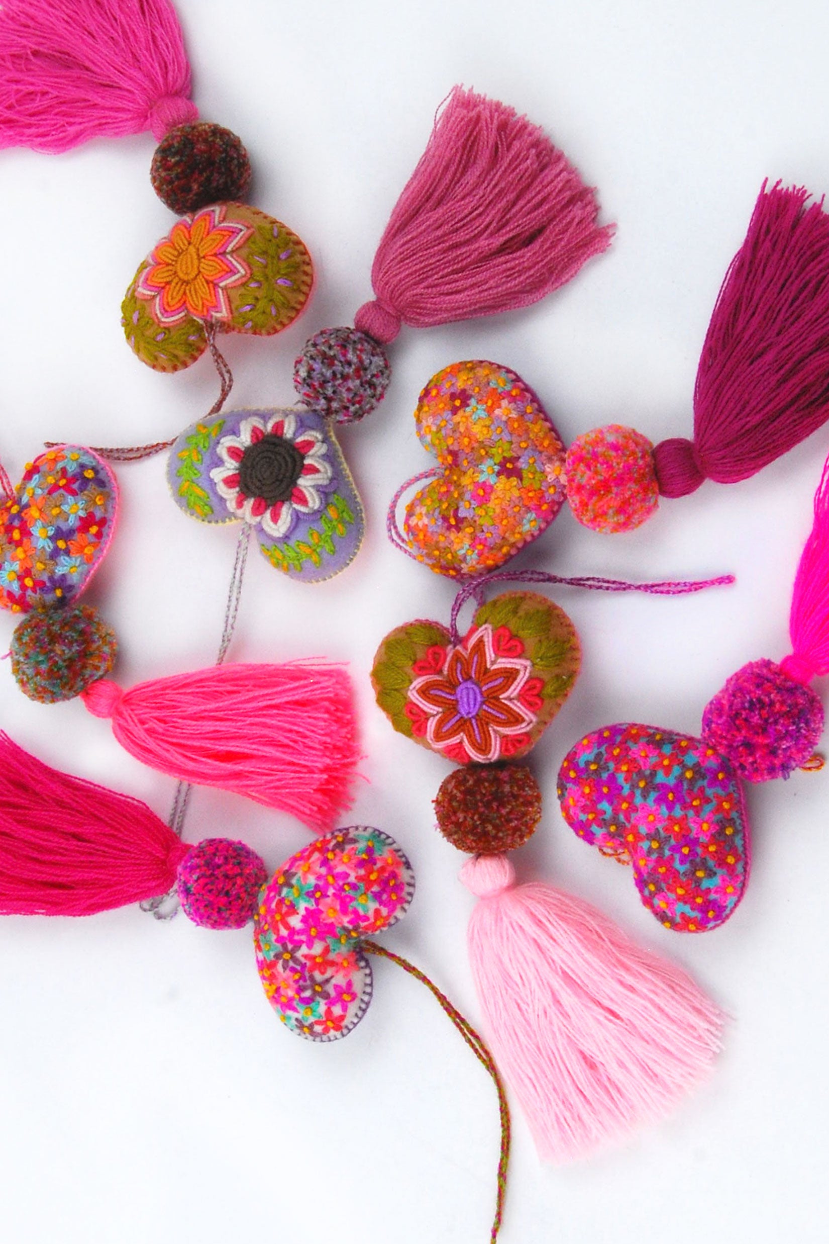 A scatter of colorful plush felt hearts adorned with flower embroidery attached by multicolor speckled pom poms to tassels in varying shades of pink. Each heart has a hanging string attached to the top of it.