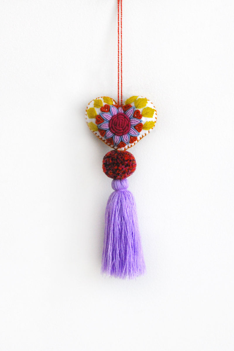 A colorful plush felt heart adorned with flower embroidery attached by multicolor speckled pom poms to a tassel in a light purple color. The heart is hanging from a string attached to the top of it.