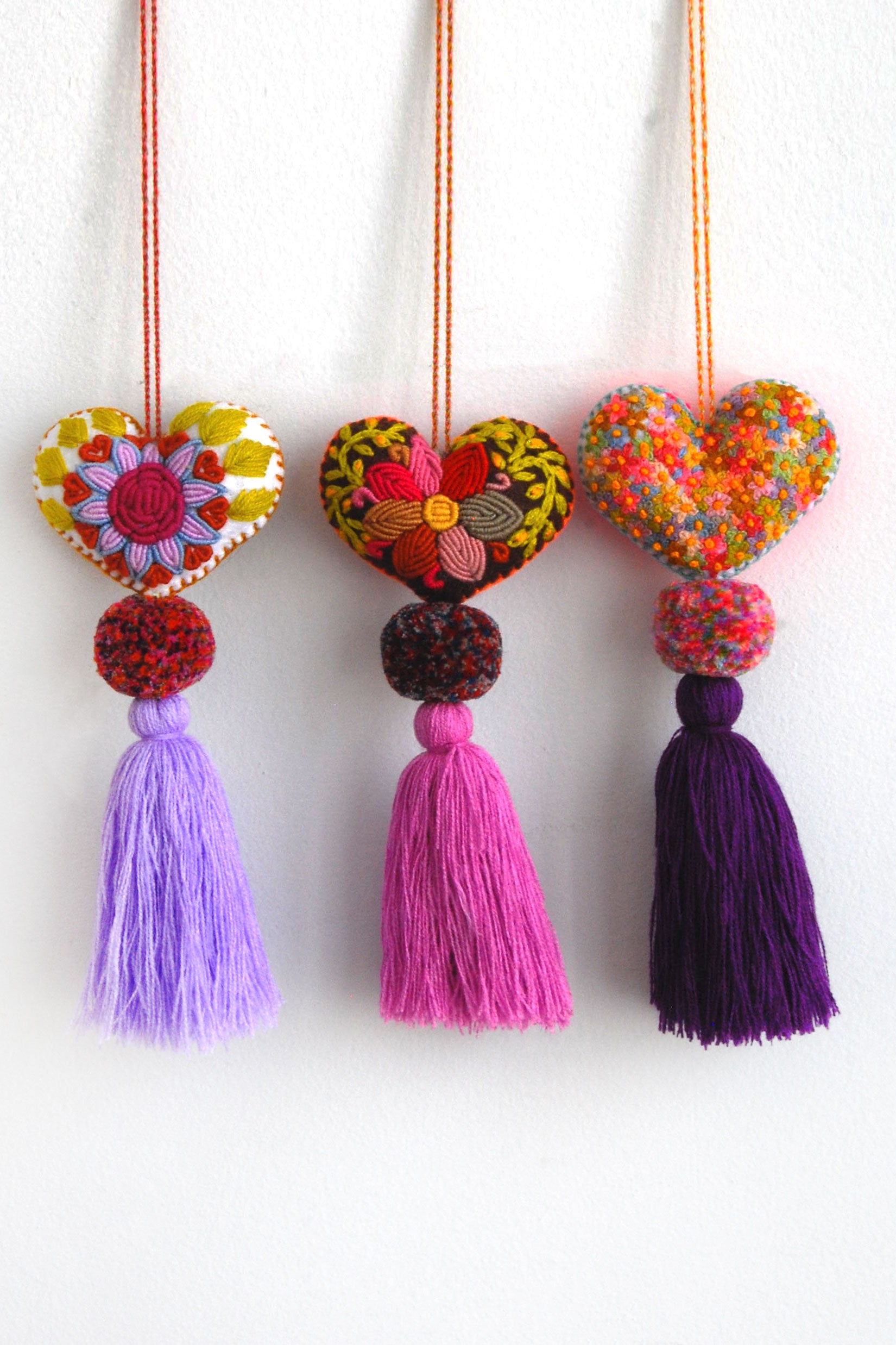 Three colorful plush felt hearts adorned with flower embroidery attached by multicolor speckled pom poms to tassels in varying shades of purple. Each heart is hanging from a string attached to the top of it.
