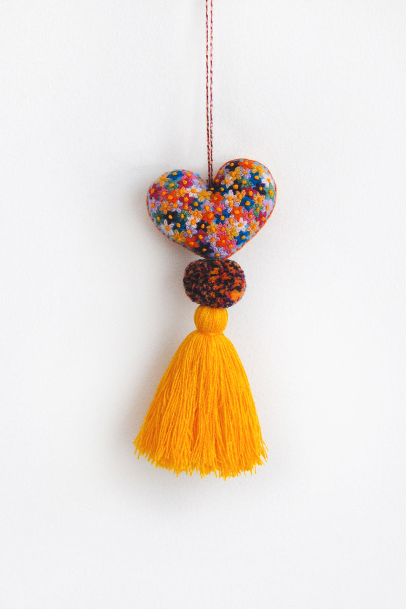 A colorful plush felt heart adorned with flower embroidery attached by multicolor speckled pom poms to a tassel in a bright gold color. The heart is hanging from a string attached to the top of it. 